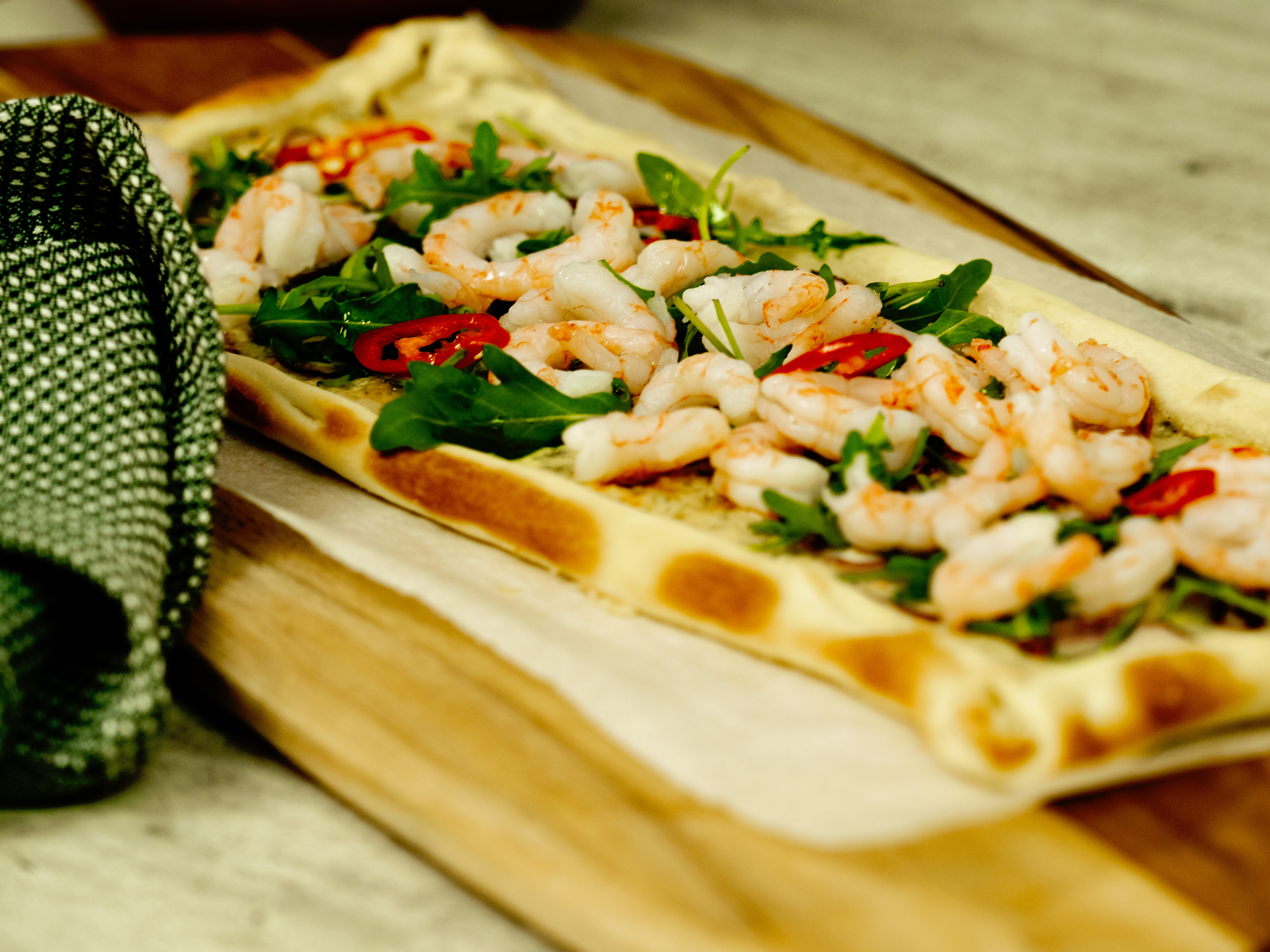 Pizza sett fra siden med reker, ruccola og chili. til høyre er et grønt kjøkkenhåndkle
