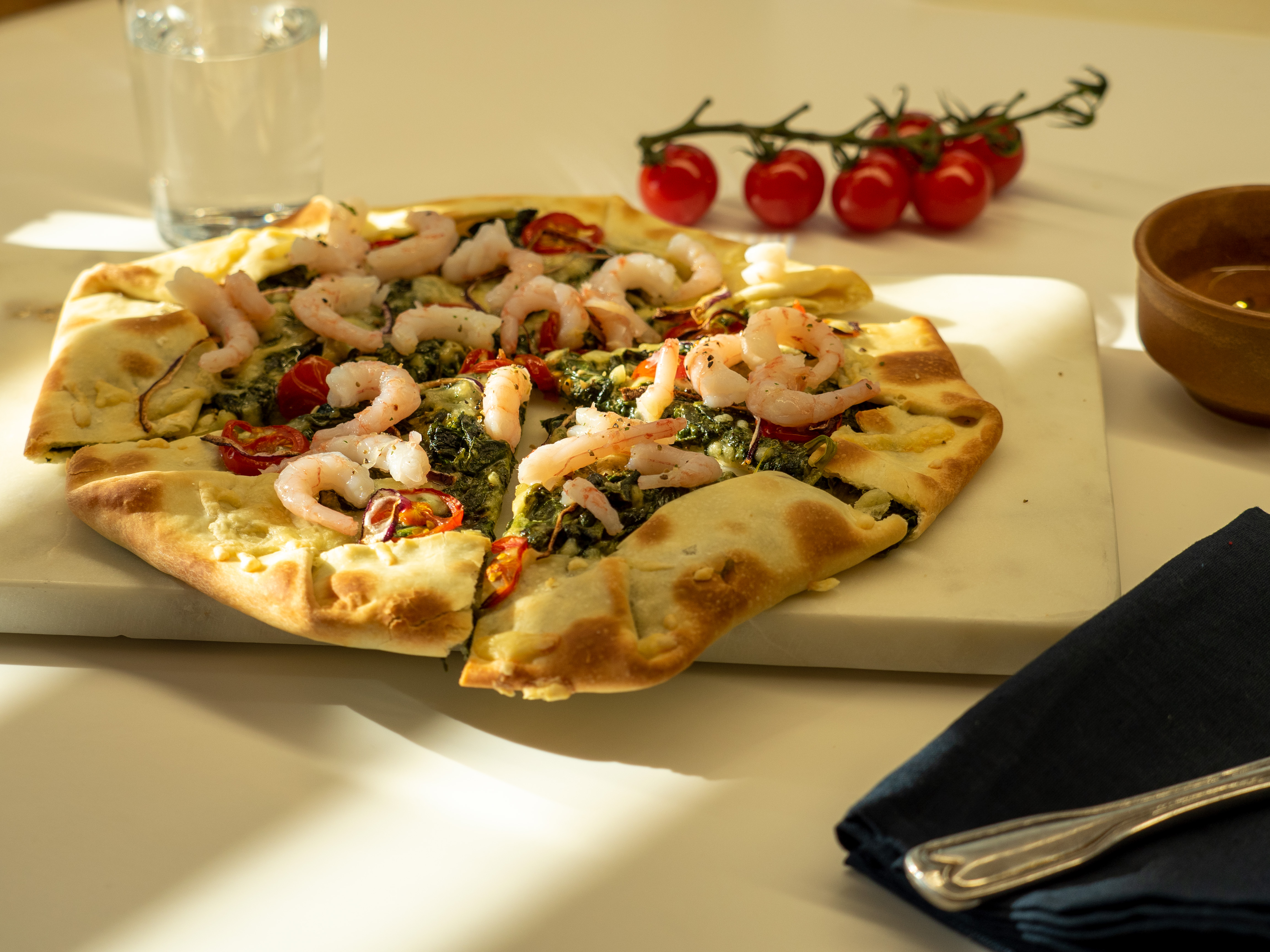 pizza sett fra siden med reker, spinat, chili, tomat. Bak er et vannglass og cherrytomater